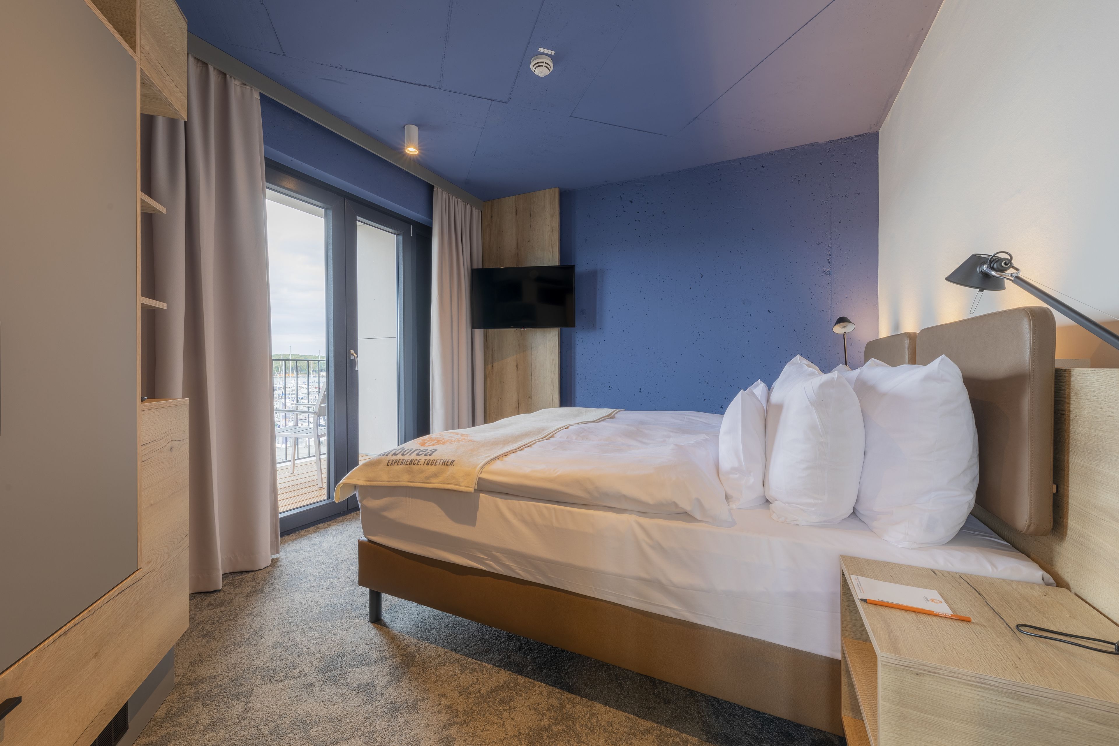 Une chambre d’hôtel avec un lit, un téléviseur fixé au mur, un bureau avec une lampe et une vue sur le balcon extérieur.