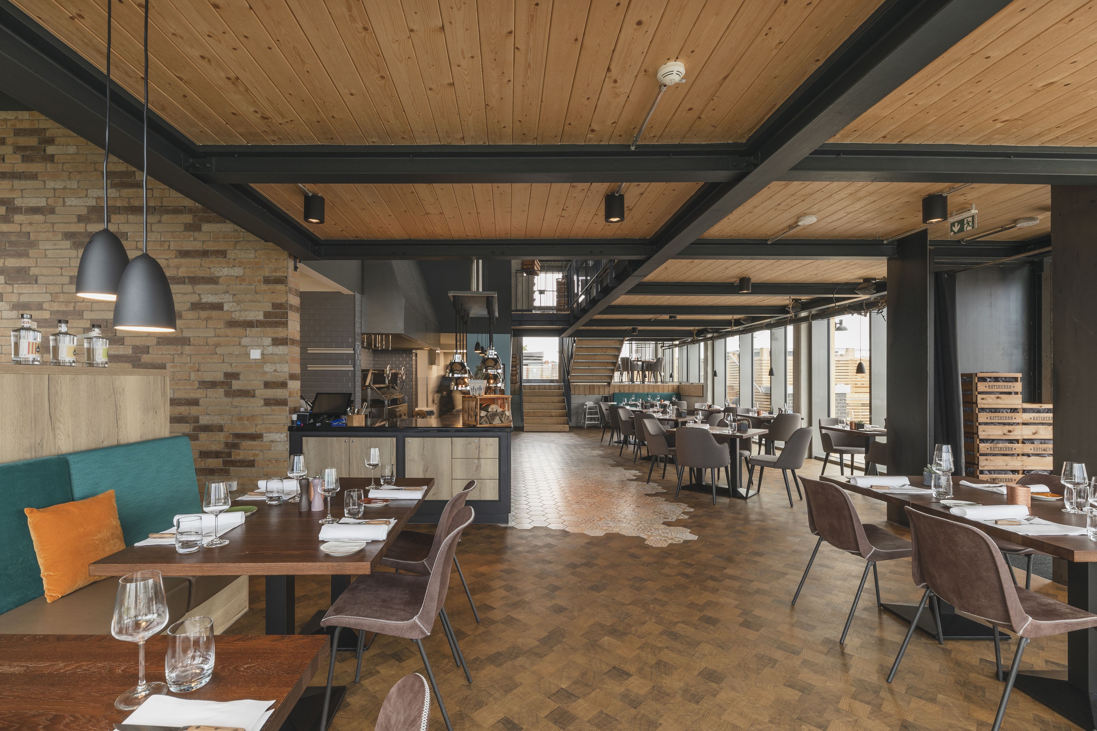 A spacious restaurant with wooden floors and high ceilings, featuring multiple dining tables and chairs, a bar area, and large windows providing natural light.