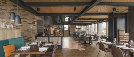 Ein geräumiges Restaurant mit Holzböden und hohen Decken, das mehrere Essplätze mit Tischen und Stühlen, einen Barbereich und große Fenster mit Tageslicht bietet.