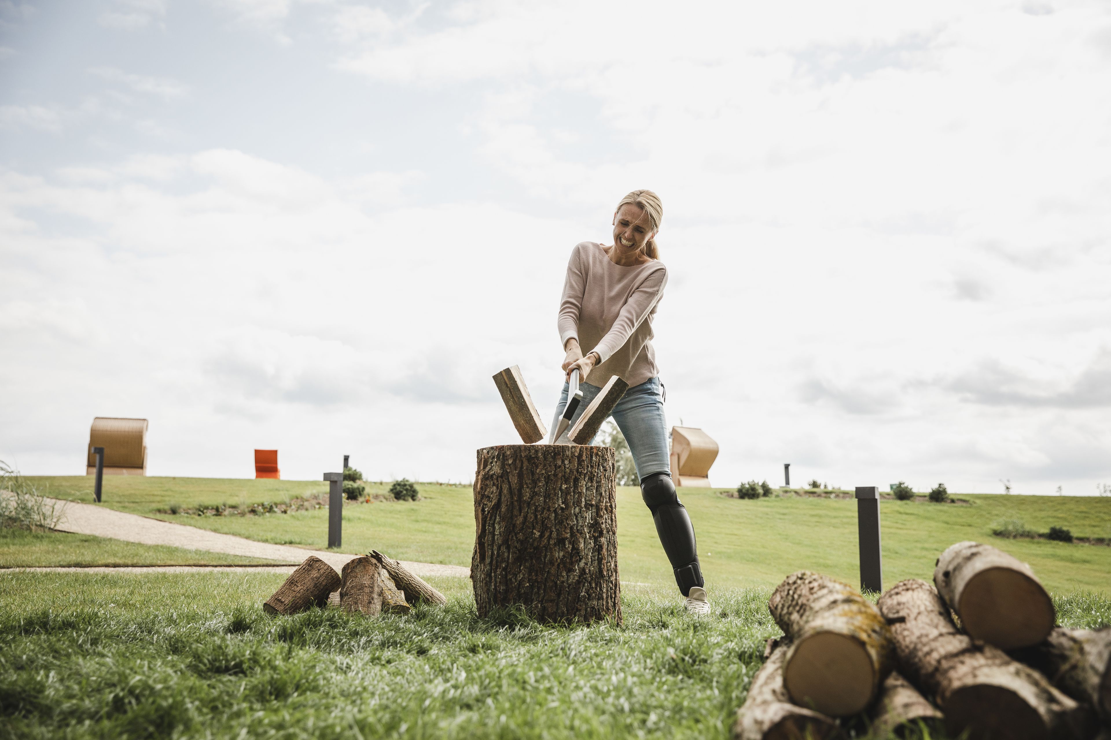A person chopping wood in a grassy field with hay bales and a tree stump in the foreground.