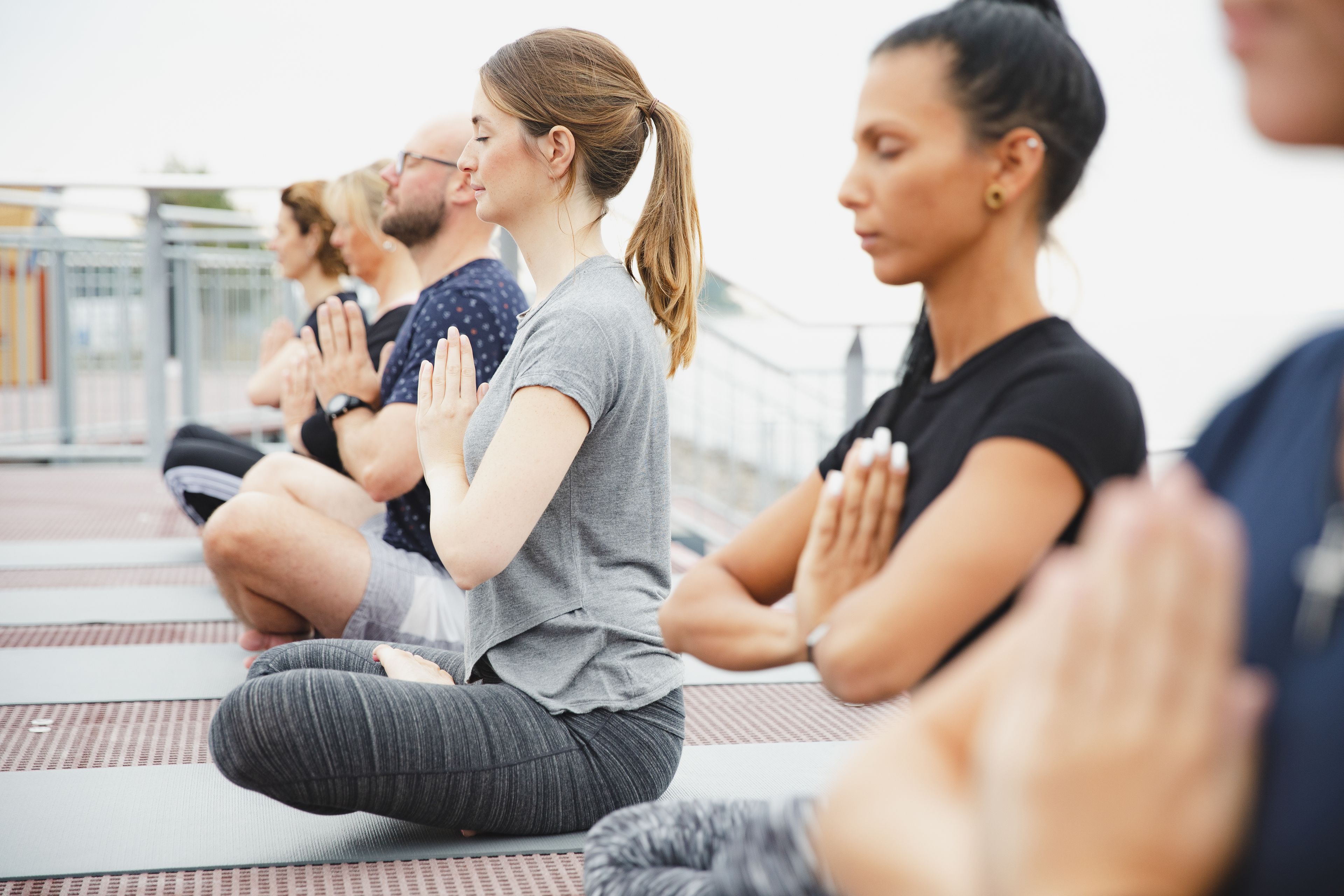 A group of people sitting on mats in a row, performing a yoga or meditation exercise outdoors.