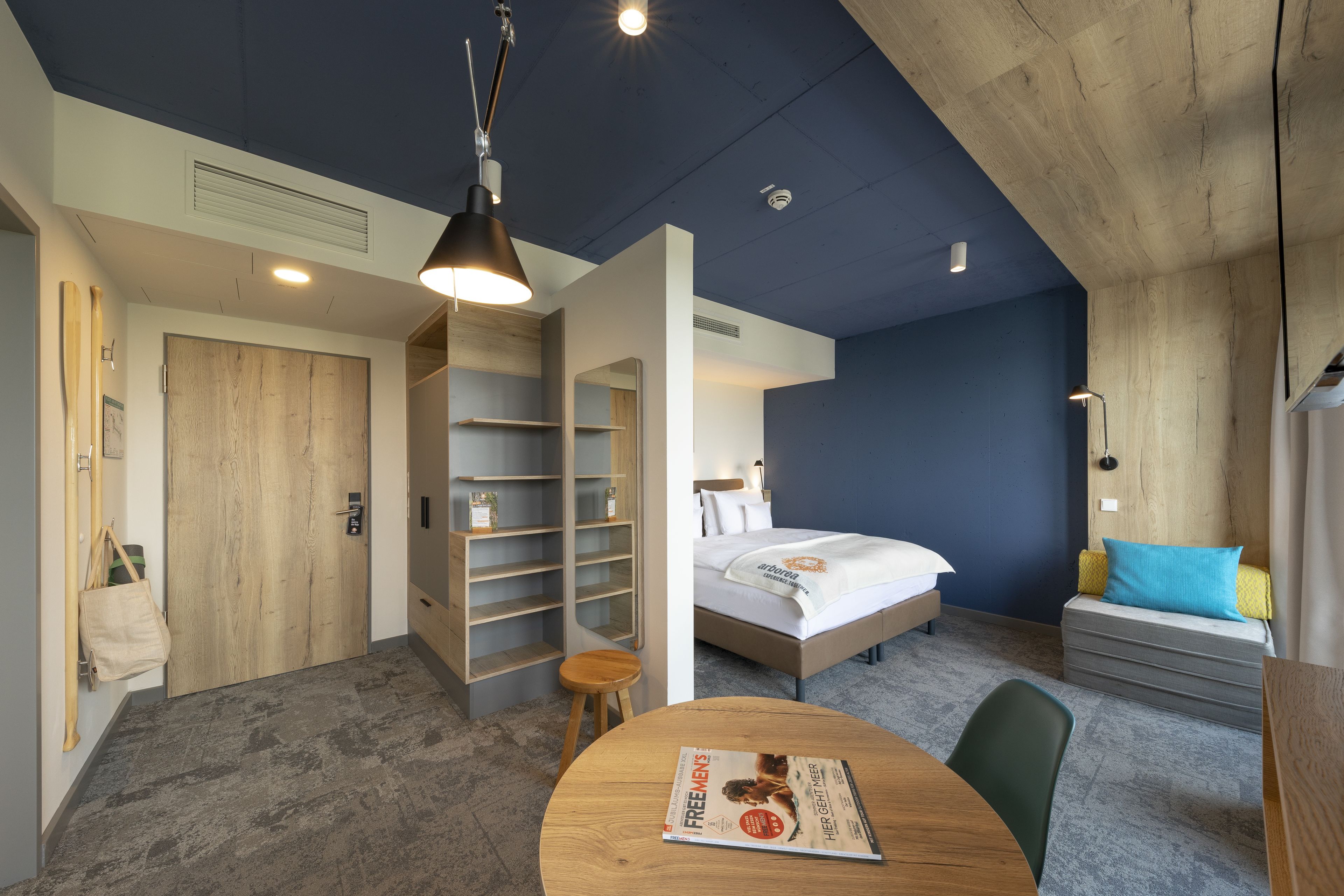 Une chambre d’hôtel moderne, équipée d’un lit, d’un bureau avec un magazine, d’une porte en bois et d’étagères intégrées.