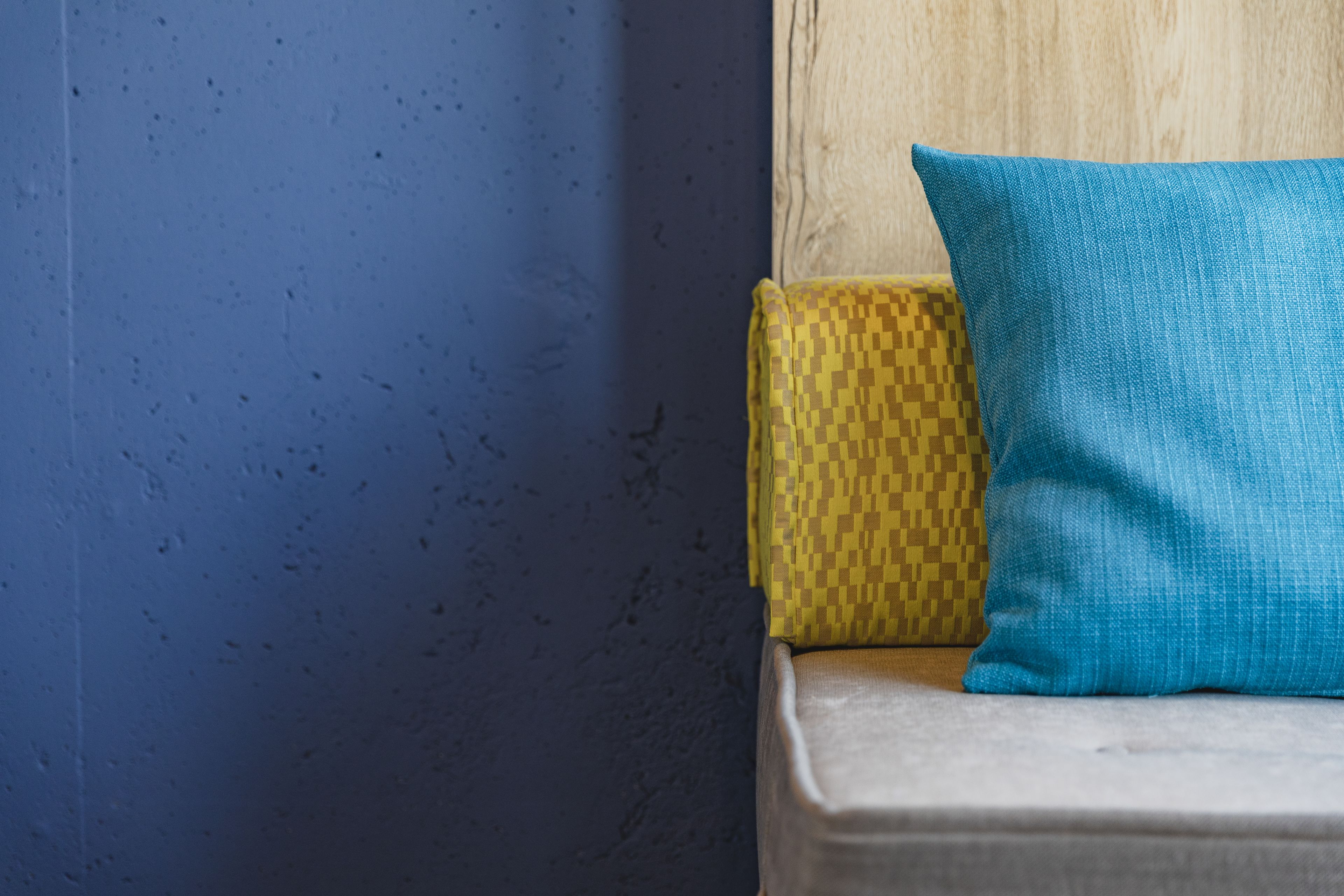 Un coin de pièce avec un mur bleu, une tête de lit en bois et deux coussins, un jaune et un bleu.