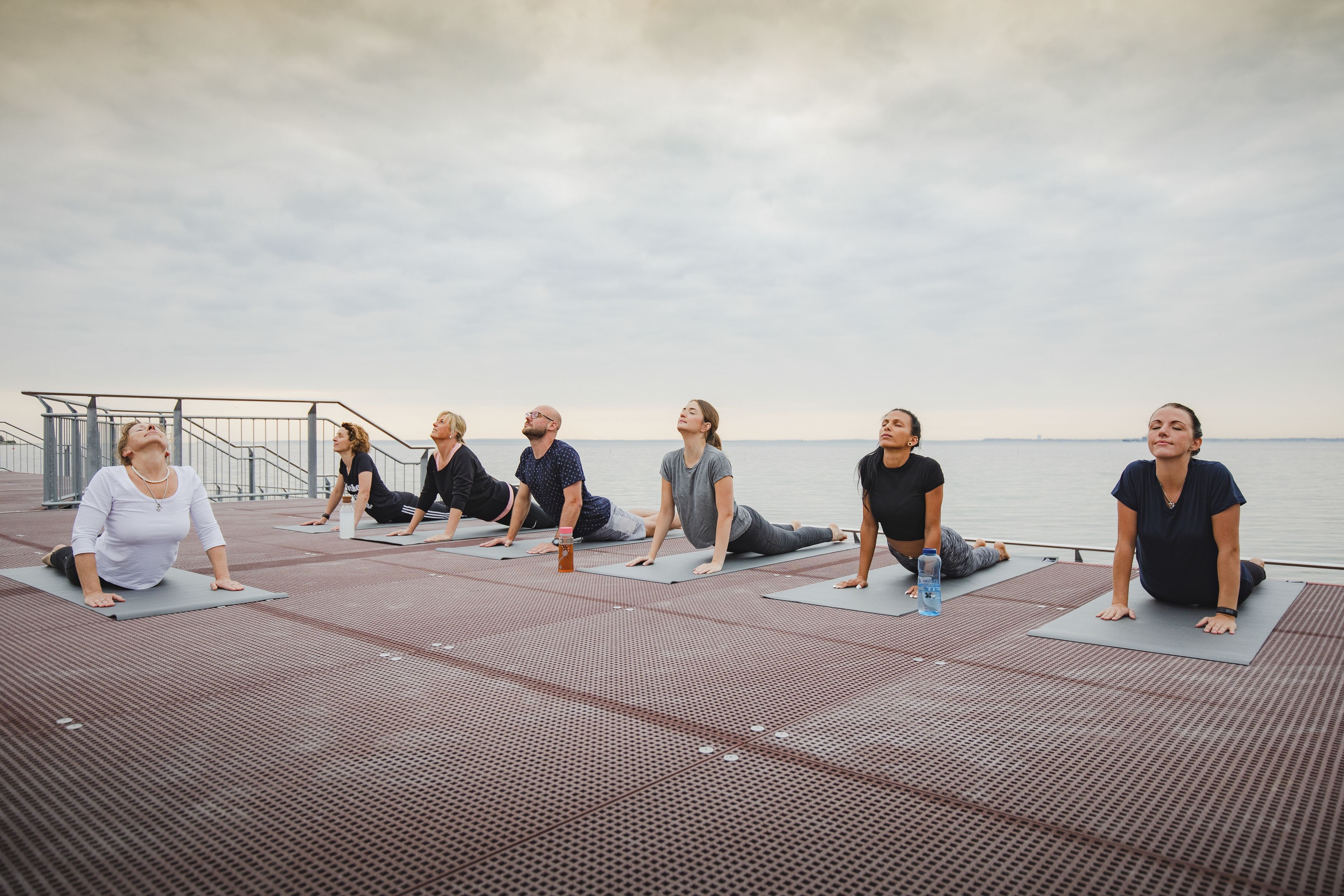 Un grupo de personas practicando yoga sobre colchonetas en un muelle, con una masa de agua al fondo.