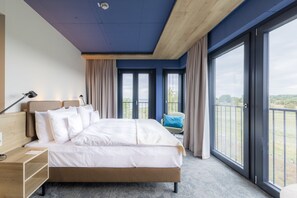 A modern hotel room with a large bed, a desk, and a view of the outdoors through large windows. - Arborea Marina Resort Neustadt (Neustadt in Holstein)