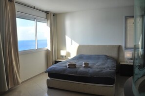 Een slaapkamer met een bed, nachtkastje, lamp en uitzicht op zee door een raam.