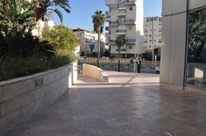A paved pathway leading to a building with a curved entrance, surrounded by palm trees and other greenery.