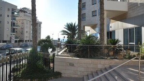 A modern building with a balcony, surrounded by palm trees and a paved area with steps leading up to the entrance. - Isra Home Luxury Apartment with Sea View (Bat Yam)