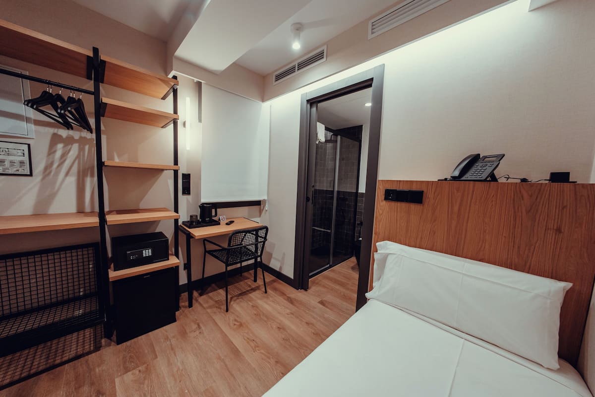 Habitación de hotel con cama, escritorio con silla, armario con estantes, espejo y teléfono sobre un soporte.