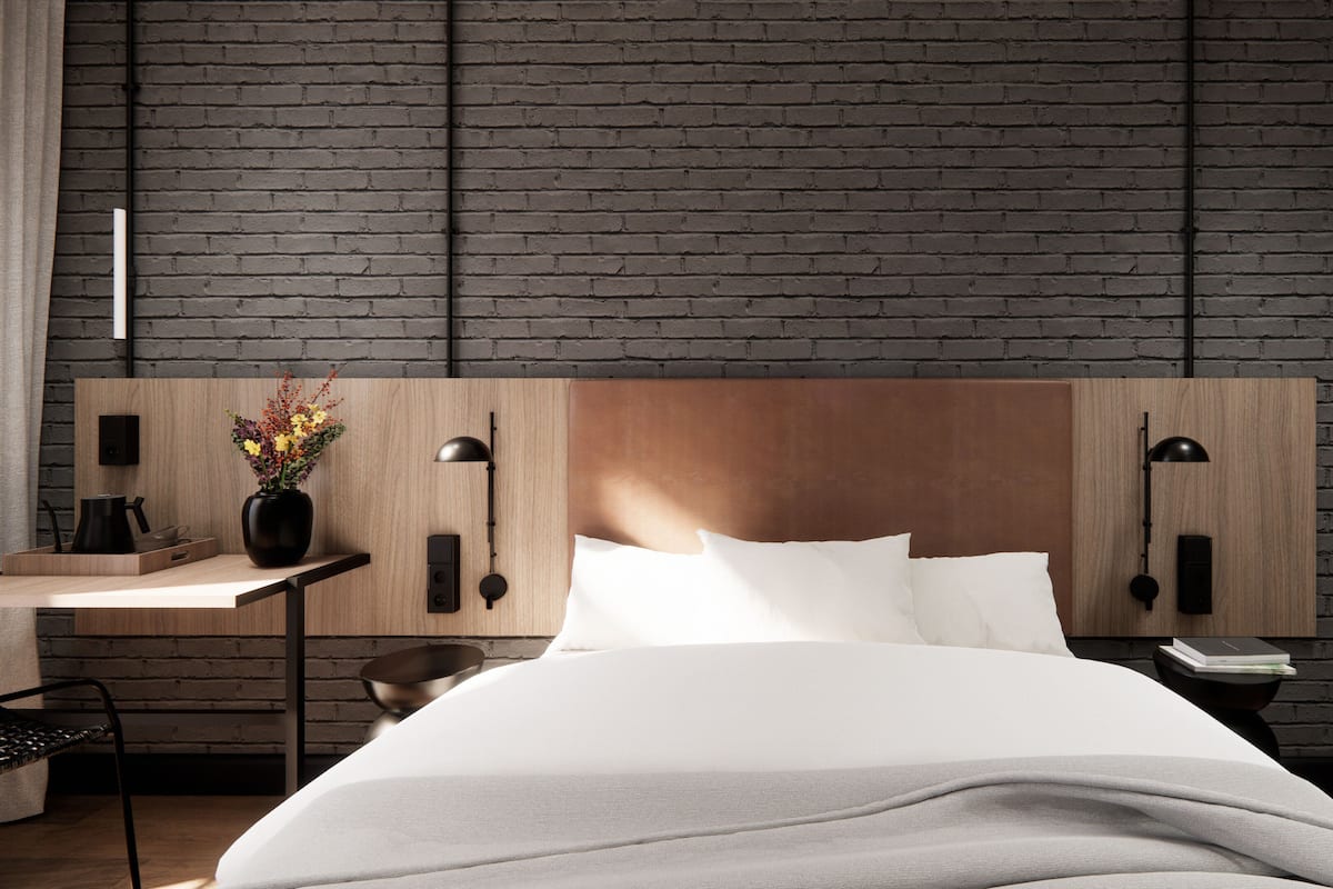 Una habitación de hotel moderna con pared de ladrillo, cabecera de madera, mesita de noche con lámpara y mesita auxiliar con un libro y un teléfono.