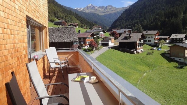 A balcony with a view of a mountain village, wooden furniture, and a table with a cup and a menu. - Appartements Veronika (Silbertal)