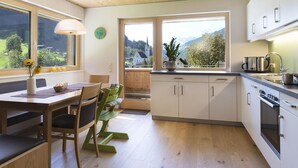 A modern kitchen with wooden cabinets, a dining area with a table and chairs, and large windows offering a view of the countryside. - Appartements Veronika (Silbertal)