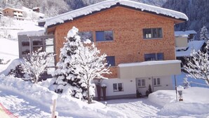 A two-story chalet with a sloped roof, surrounded by snow-covered trees and mountains in the background. - Appartements Veronika (Silbertal)