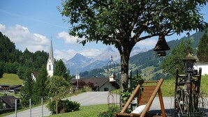 A rural setting with a wooden swing chair, a white church, and a mountain backdrop. - Appartements Veronika (Silbertal)