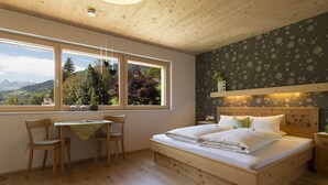 A bedroom with a wooden bed, a desk with chairs, a window with a view of mountains, and a floral wallpaper. - Appartements Veronika (Silbertal)