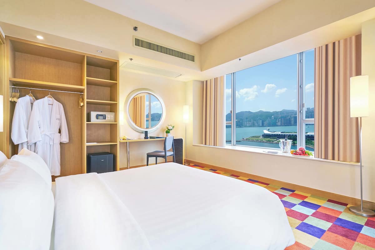 Un dormitorio de hotel con una cama grande, un armario, un escritorio con espejo, vistas a un paisaje urbano a través de un ventanal amplio y una alfombra de colores.