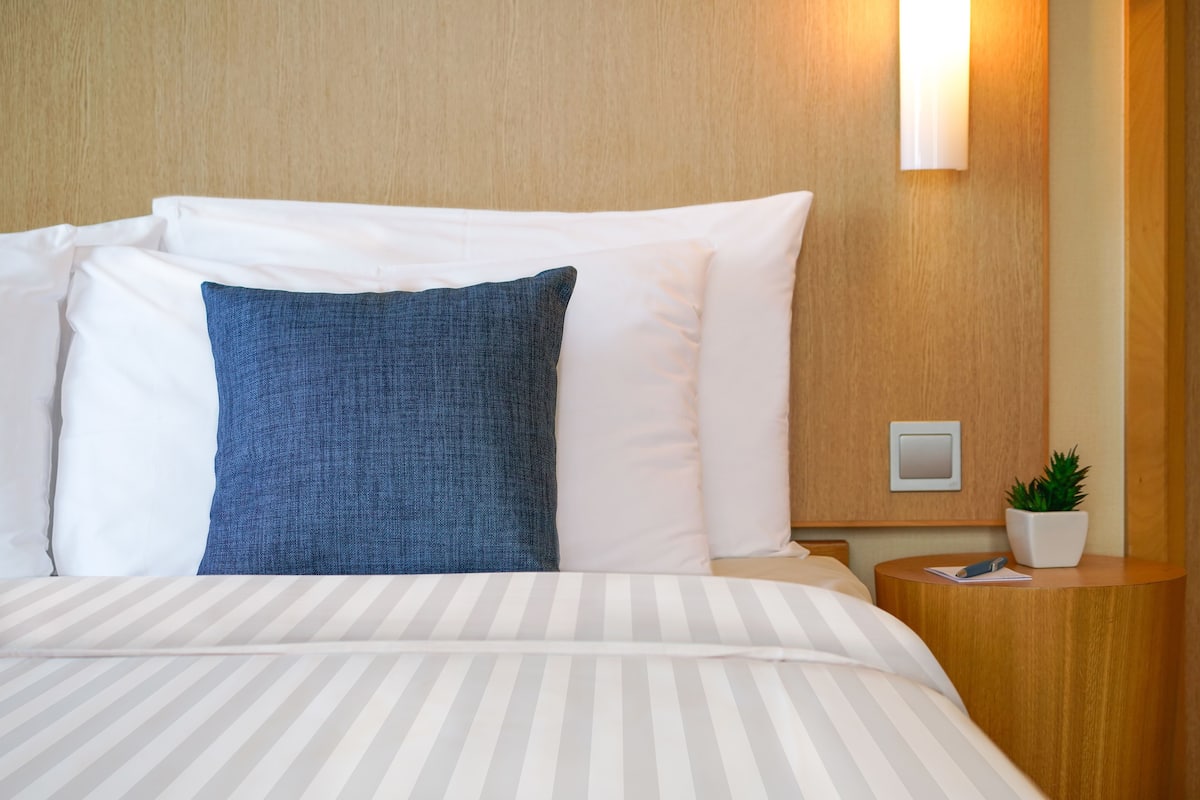 Una cama bien hecha con almohadas blancas y azules, una colcha a rayas, una mesita de noche con una planta y una lámpara de pared.