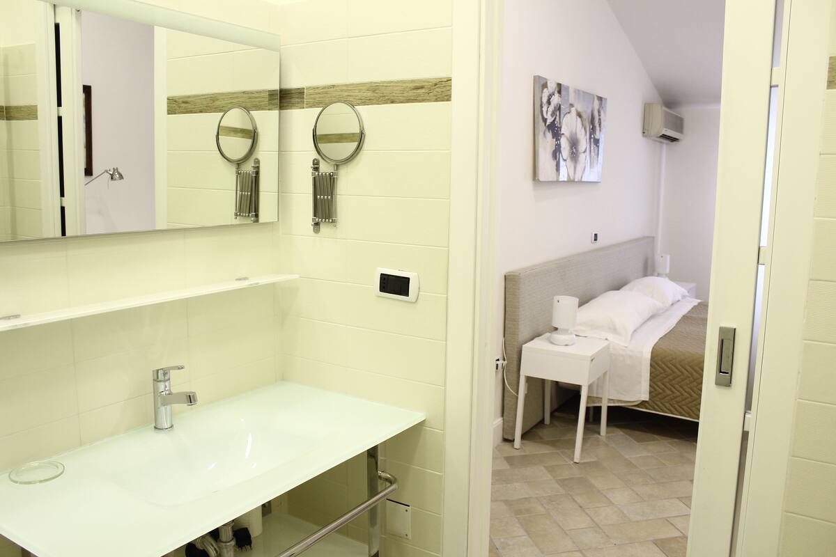 Ein Badezimmer mit einem weißen Waschbecken, zwei Wandspiegeln, einem Handtuchhalter und einem Bett mit weißem Kopfteil.