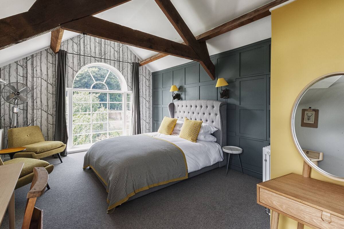 A bedroom with a large bed, a wooden desk, a chair, a round mirror, and a window with a view of greenery.