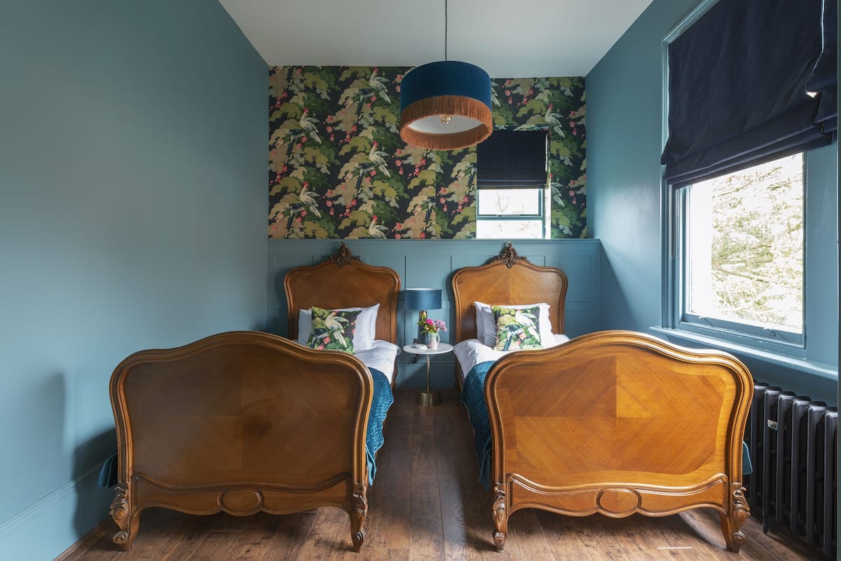 A room with two wooden beds, a blue wall, a floral wallpaper, a round table with a lamp, and a window with a view.