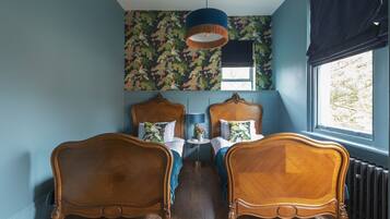 A room with two wooden beds, a blue wall, a floral wallpaper, a round table with a lamp, and a window with a view.