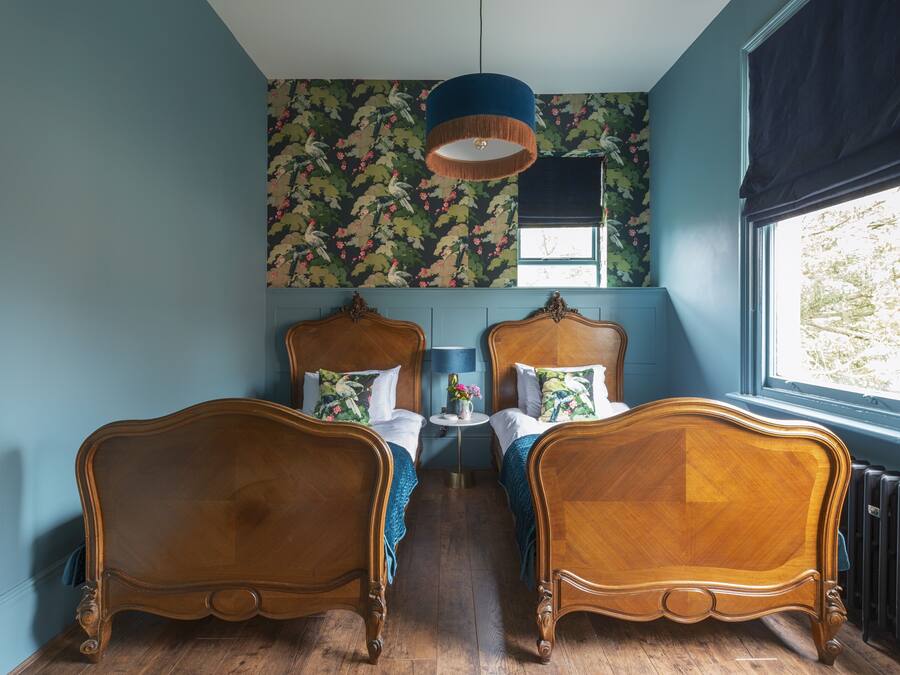 A room with two wooden beds, a blue wall, a floral wallpaper, a round table with a lamp, and a window with a view.