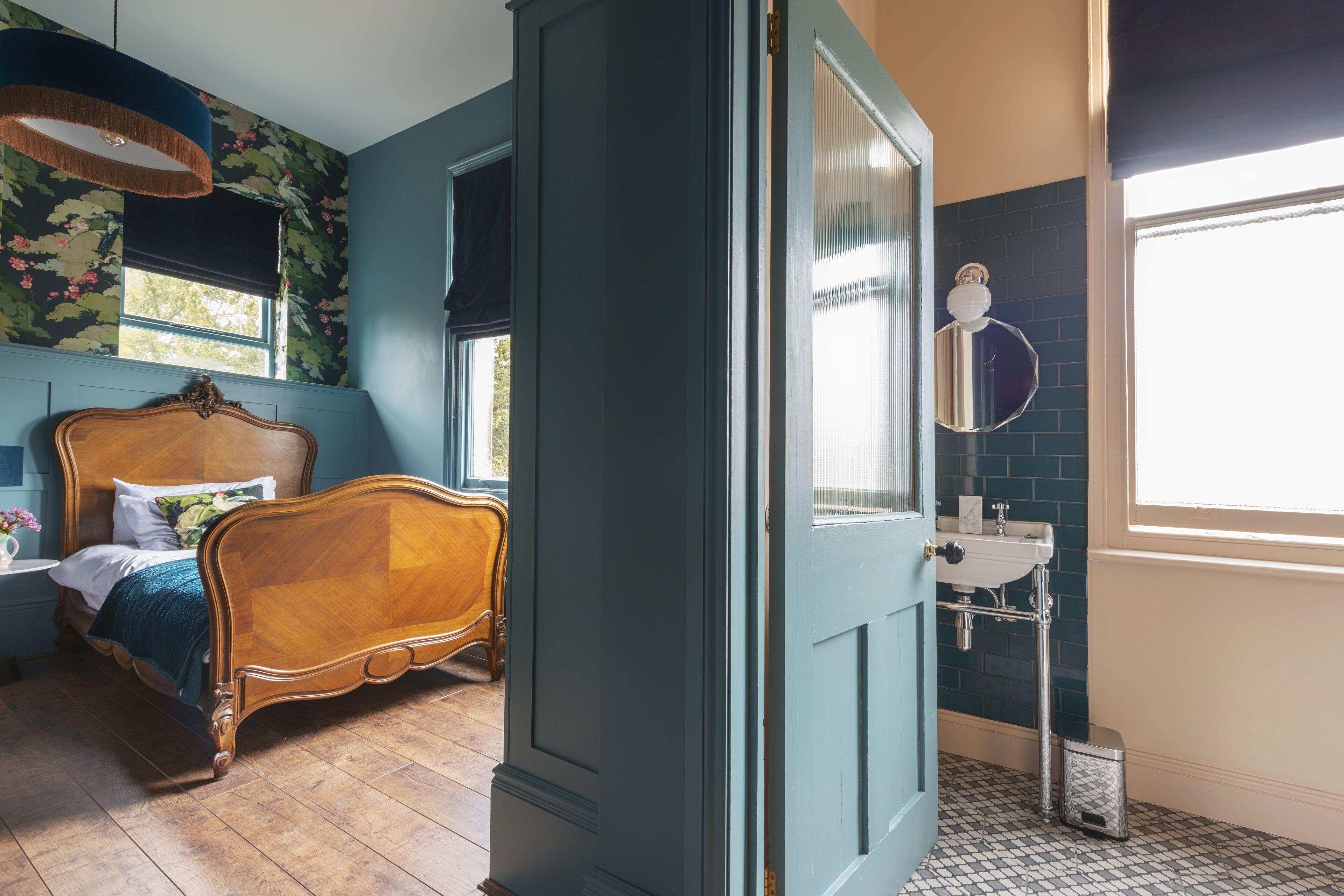 Un dormitorio con cama de madera, ventana con vistas, baño con lavamanos y espejo, y una puerta azul.