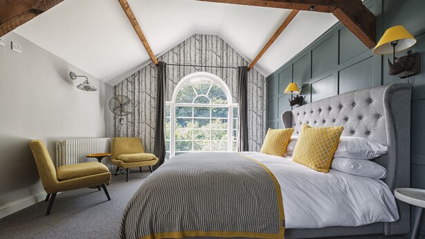 A bedroom with a large bed, two chairs, a small table, and a window with a view of greenery. - The Coach House (Derby)
