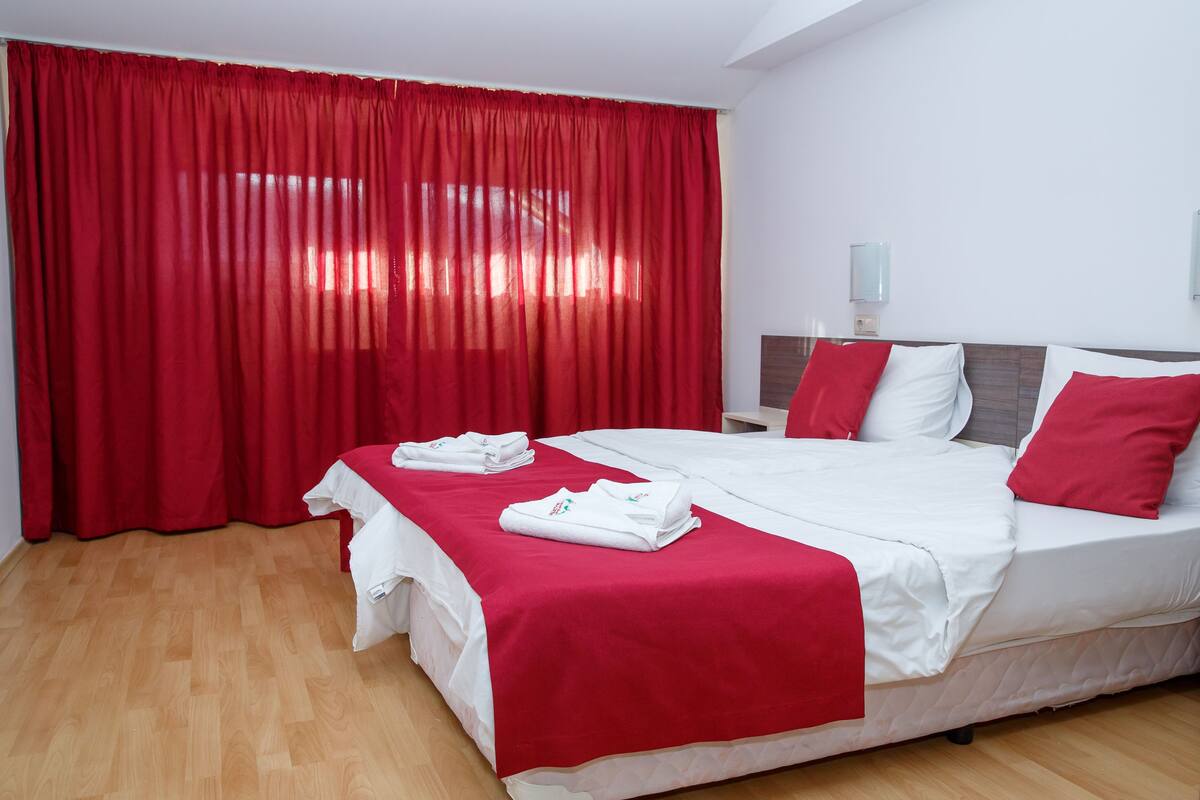 A hotel room with a large bed, red curtains, white bedding, and a wooden floor.