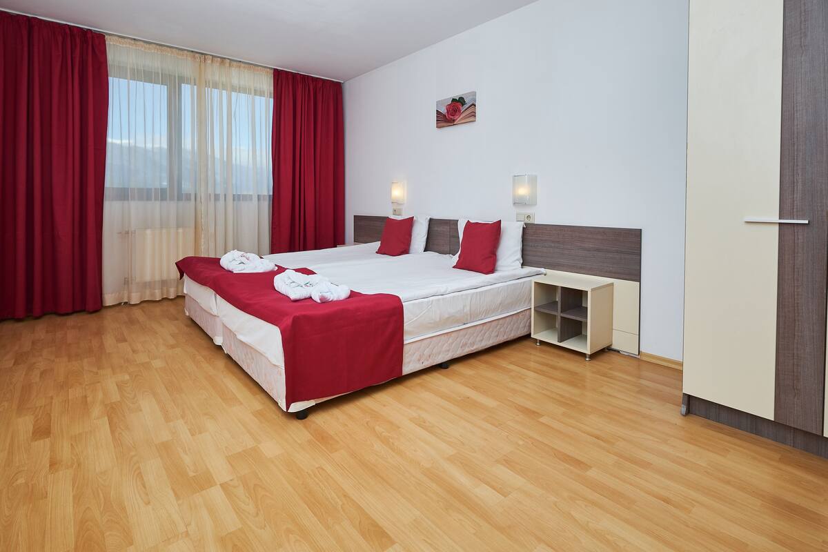 A hotel room with a large bed, red curtains, a wooden floor, a small bedside table with a lamp, and a wall-mounted artwork.