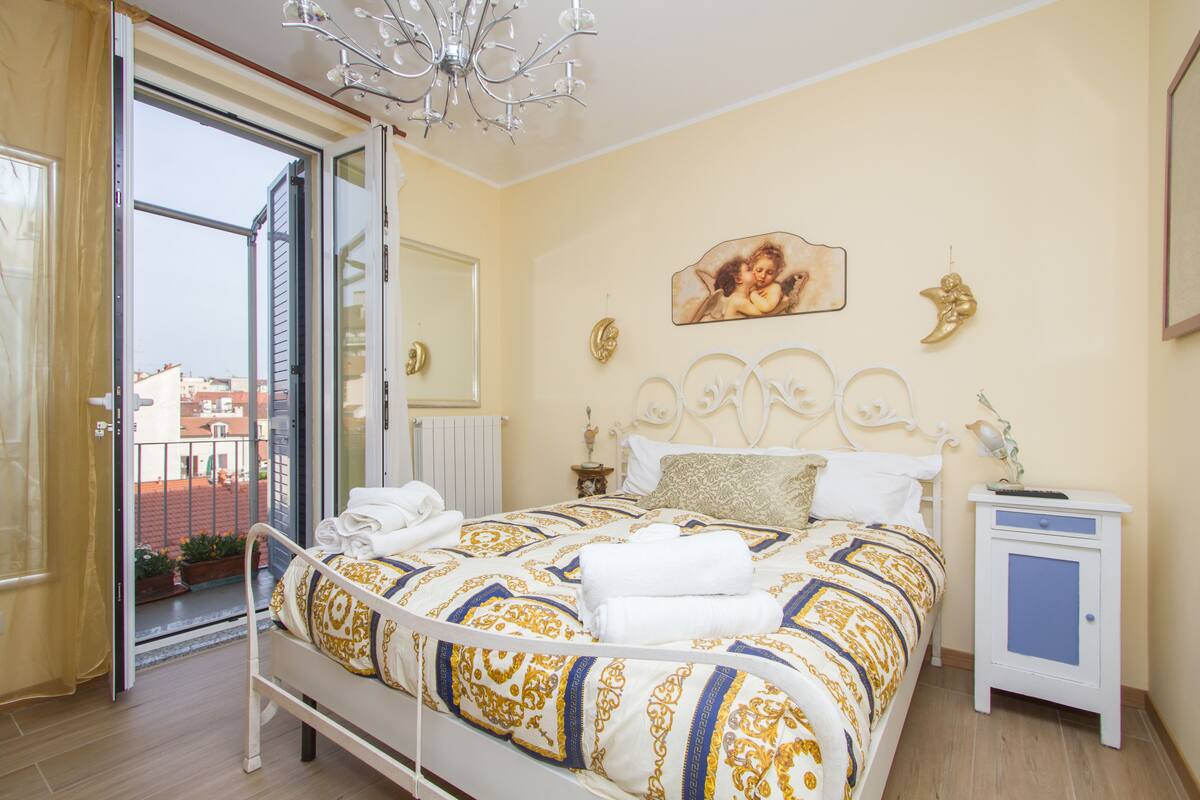 Ein Schlafzimmer mit einem Bett, einem Nachttisch, einer Chandelierleuchte, einem Fenster und Blick auf Gebäude.