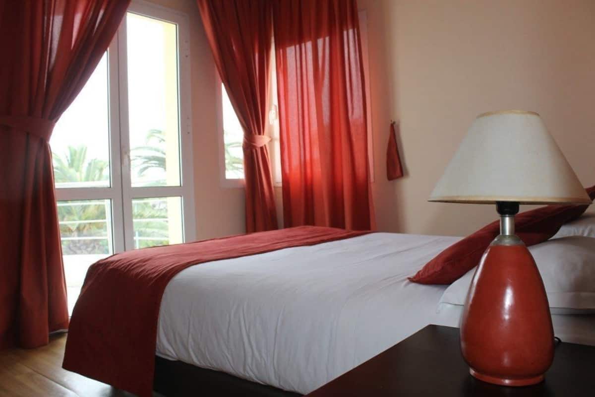 Habitación de hotel con cama, cortinas rojas, lámpara blanca y vista a palmeras a través de una ventana.