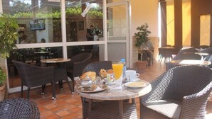 Uma área de estar externa com móveis de vime, uma mesa com itens de café da manhã e vista do interior através de portas de vidro.