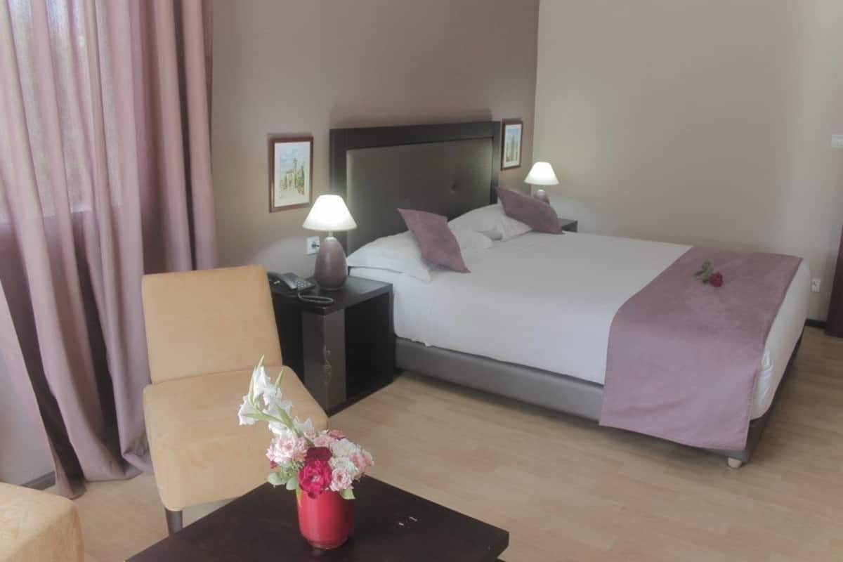 Habitación de hotel con cama, mesitas de noche, una silla y un florero con flores.
