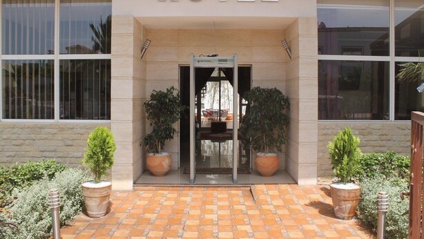 Entrada de hotel com um pátio ladrilhado, plantas em vasos e uma placa com a inscrição 'HOTEL'.