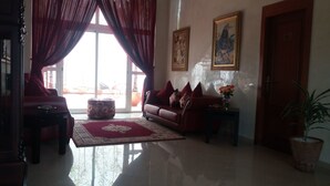 Sala de estar com sofá, banquinho e mesa de centro. Há uma porta e uma janela com cortinas.