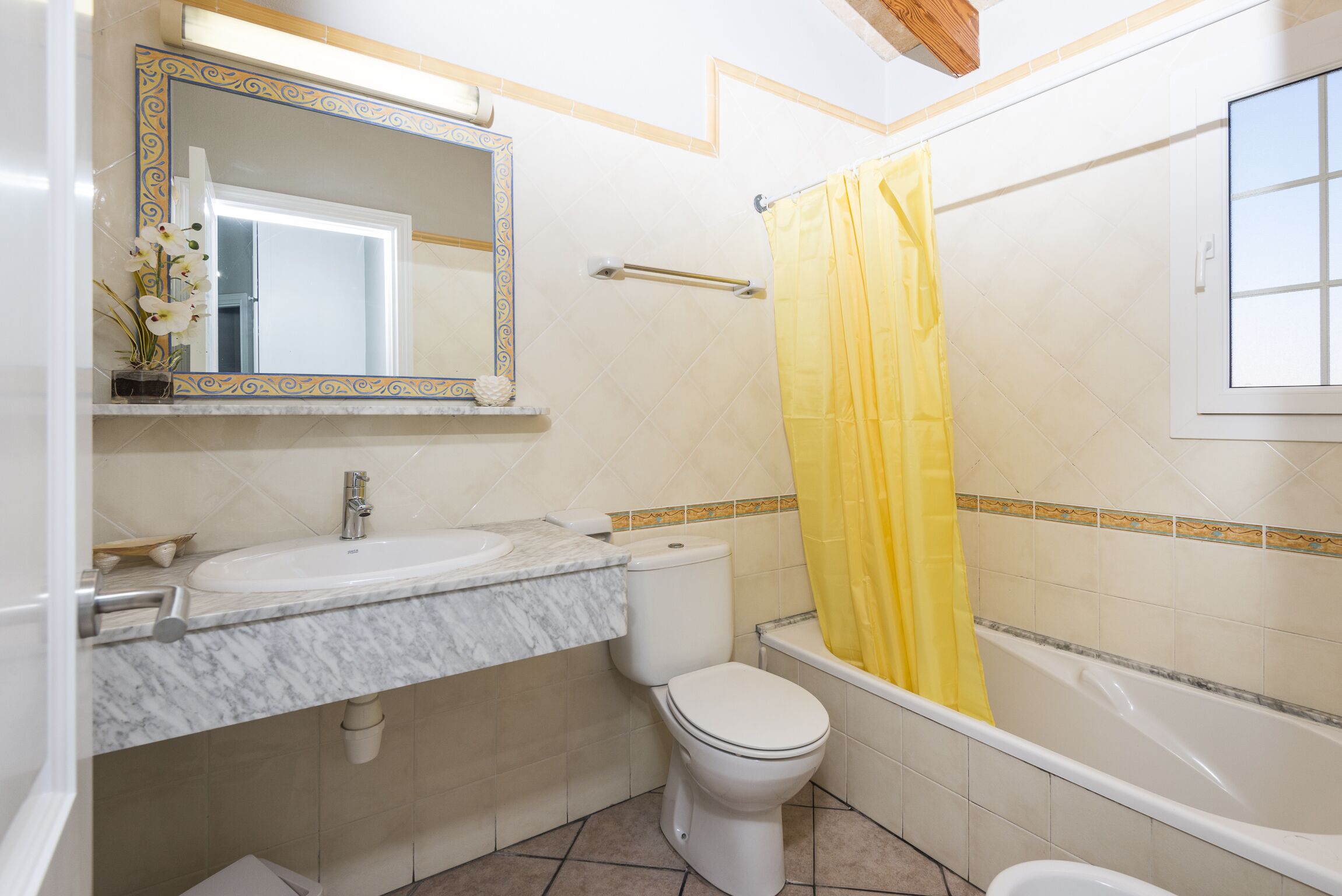 Een badkamer met een wit toilet, een wastafel met een marmeren blad, een badkuip met een geel douchegordijn en een spiegel met een decoratieve lijst.