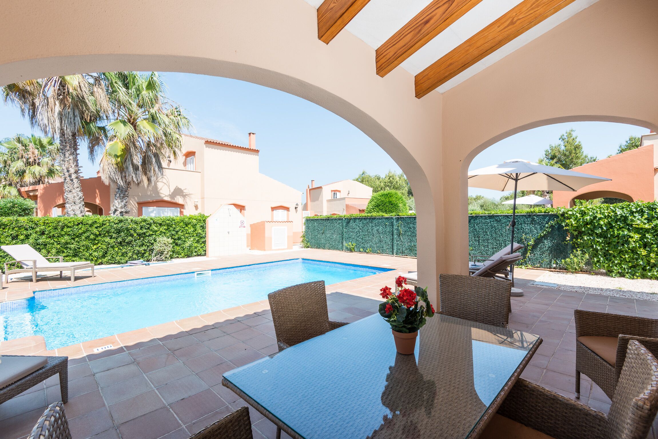 A poolside area with a glass table and wicker chairs, surrounded by a pool and palm trees.