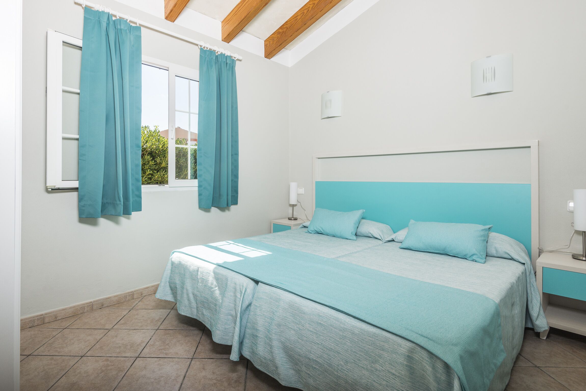 A bedroom with a bed, a nightstand, a window with curtains, and a ceiling with wooden beams.