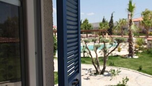 A view through a window with horizontal blinds, a swimming pool, palm trees, and a building with a red roof in the background. - Igdeli Han Hotel (Cesme)