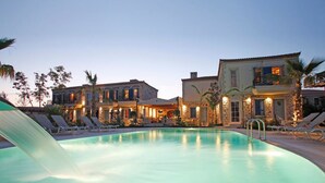 A swimming pool with lounge chairs and a water slide, surrounded by buildings with stone facades and warm lighting. - Igdeli Han Hotel (Cesme)
