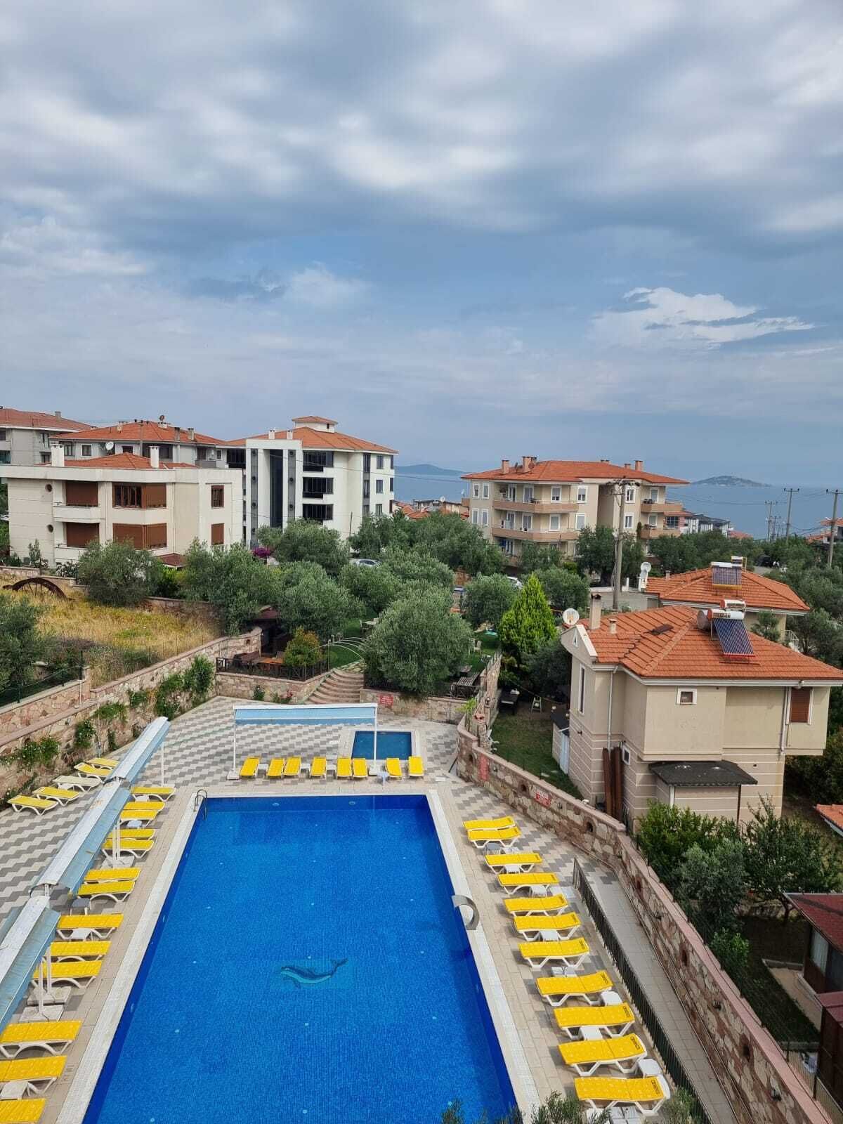 A swimming pool with yellow lounge chairs, surrounded by buildings with red roofs and a view of the sea.