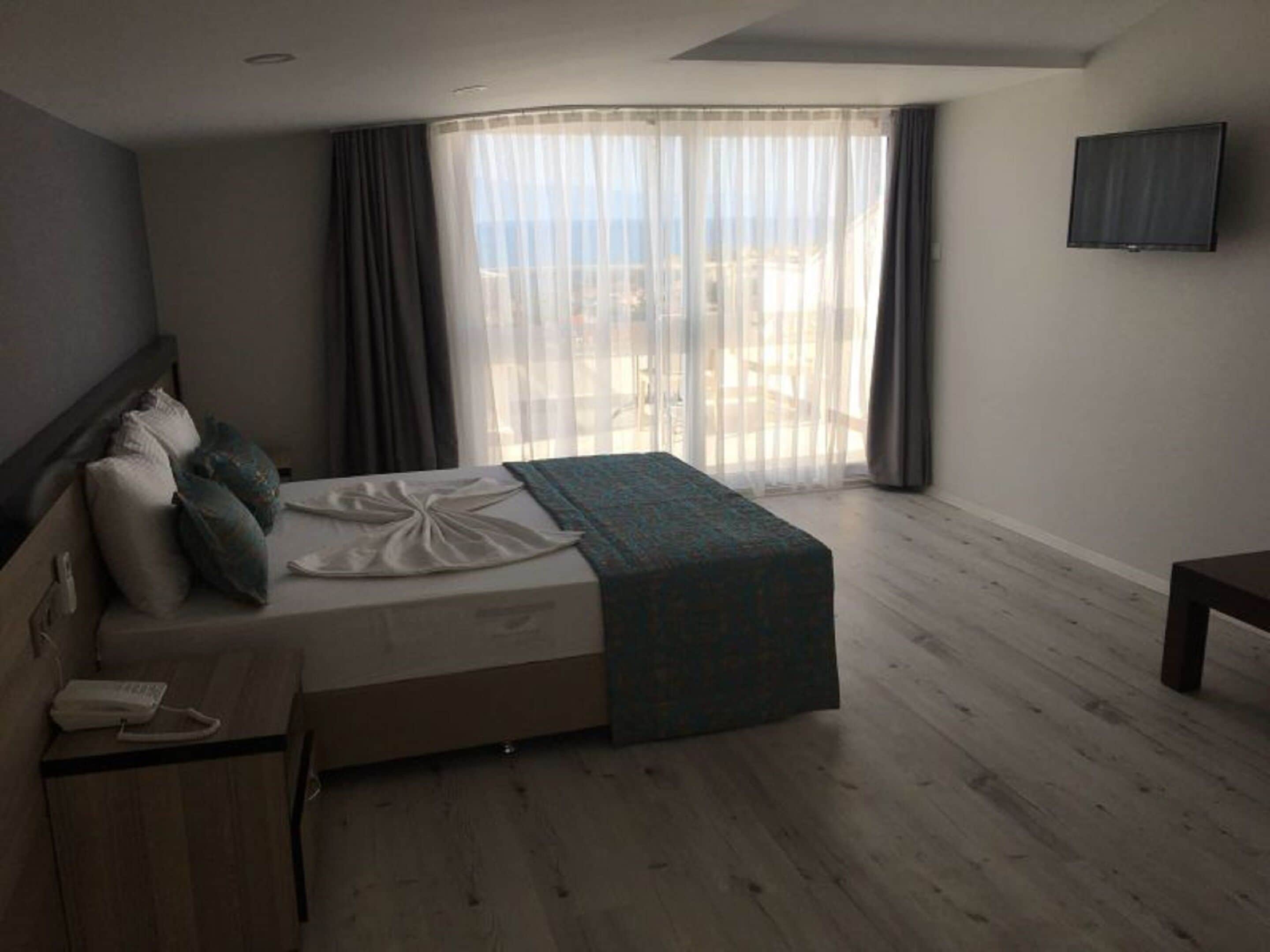 A hotel room with a bed, a bedside table with a phone, a TV on the wall, and a large window with curtains.