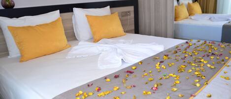 Una cama bien hecha con almohadas amarillas y blancas, una toalla blanca y pétalos de flores esparcidos.