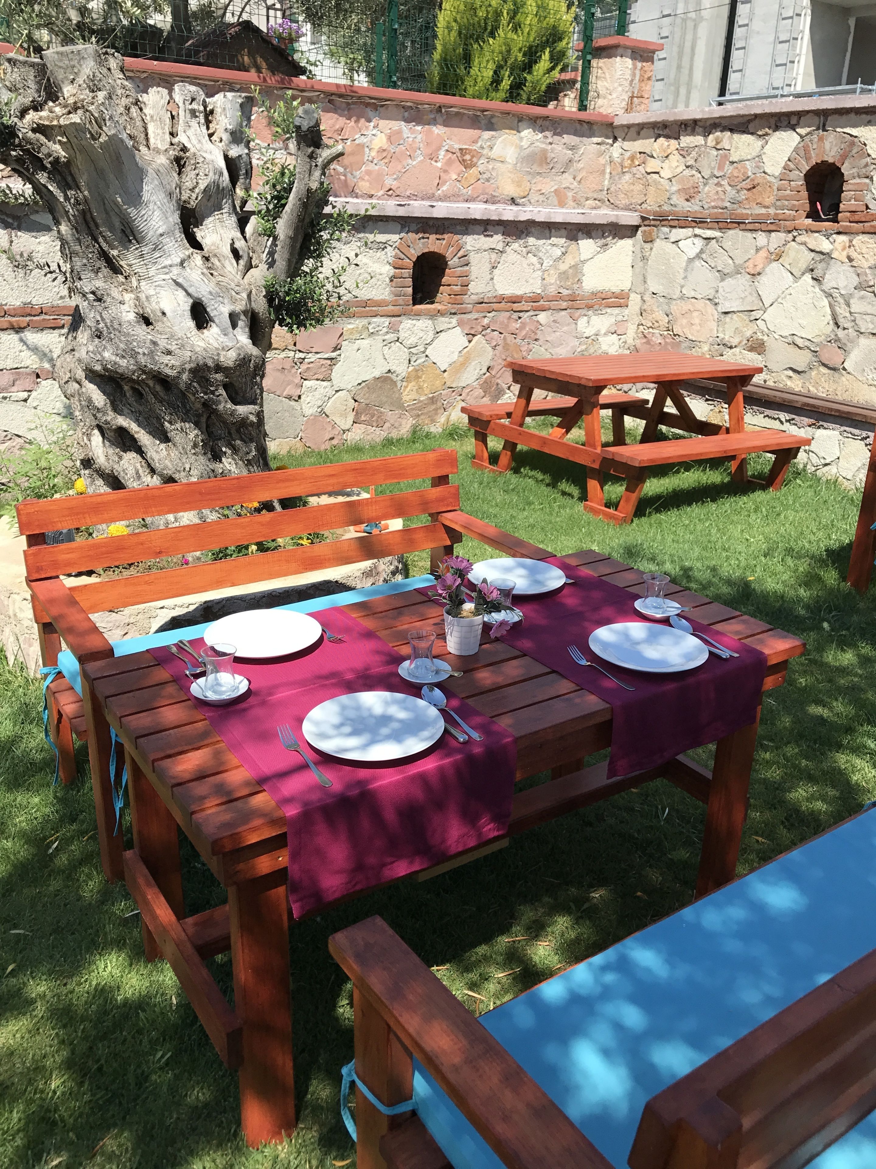 Een houten picknicktafel gedekt met borden, bestek en een glazen pitcher, omringd door een stenen muur en een grote boom.