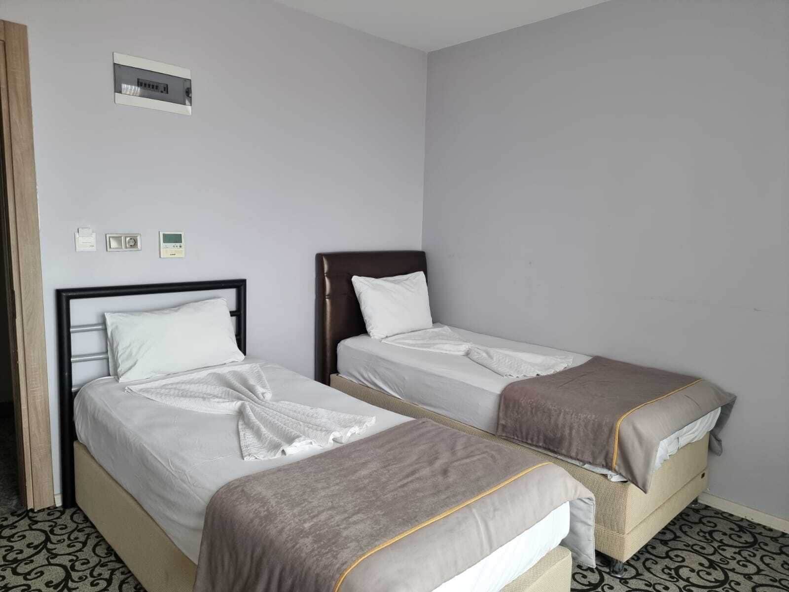 A hotel room with two single beds, a small bedside table, a wall-mounted power outlet, and a light switch.