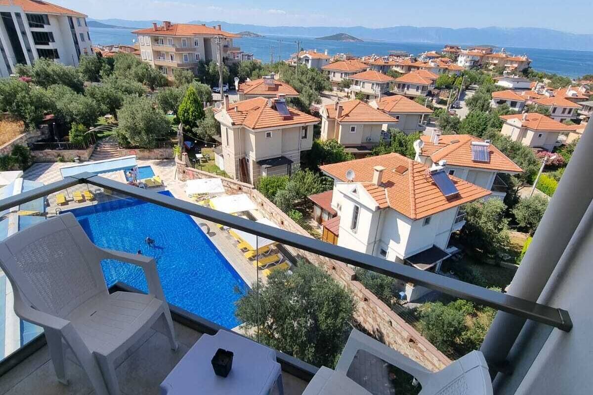A balcony with a pool, white lounge chairs, and a view of a residential area with red-roofed houses and a body of water in the distance.