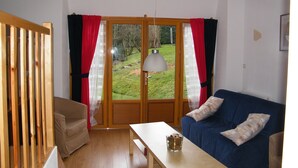 A room with a wooden floor, a blue sofa, a wooden table, and a view of a garden through a glass door. - Maison Neuve Grandval - Gite (Grandval)