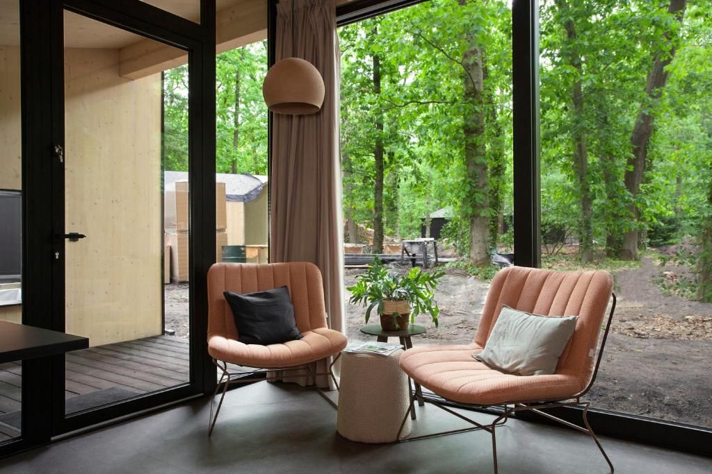 A room with two cushioned chairs, a small round table, and a view of a forest through large windows.