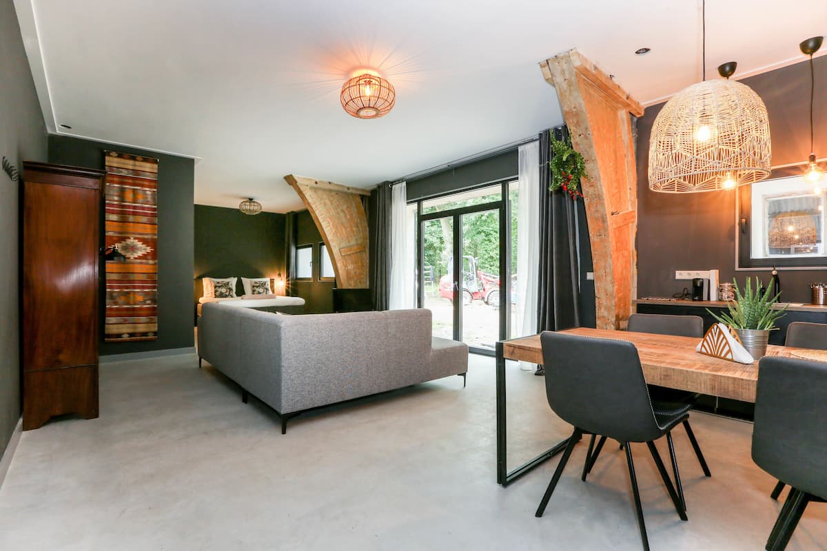 Una sala moderna con sofá gris, mesa de comedor de madera y sillas negras. Hay un ventanal con cortinas, un mueble de madera y una lámpara colgante.